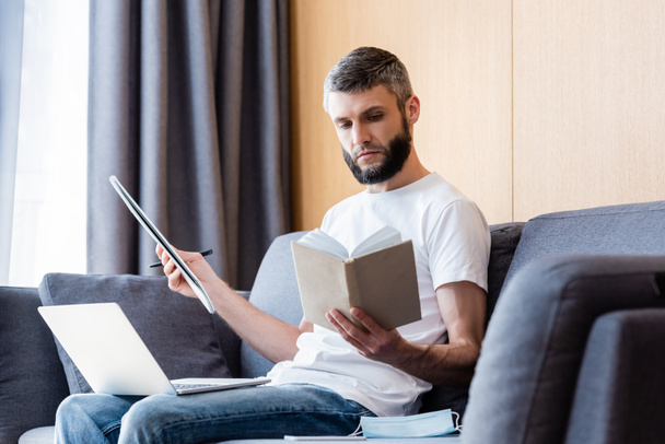 Вибірковий фокус книги для читання телефону під час роботи з ноутбуком та ноутбуком біля медичної маски на дивані
  - Фото, зображення