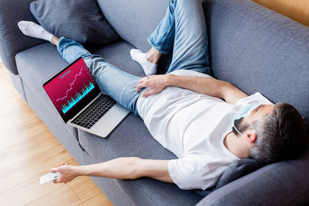Человек в медицинской маске держит смартфон во время сна на диване рядом с ноутбуком с графиками на экране
  - Фото, изображение