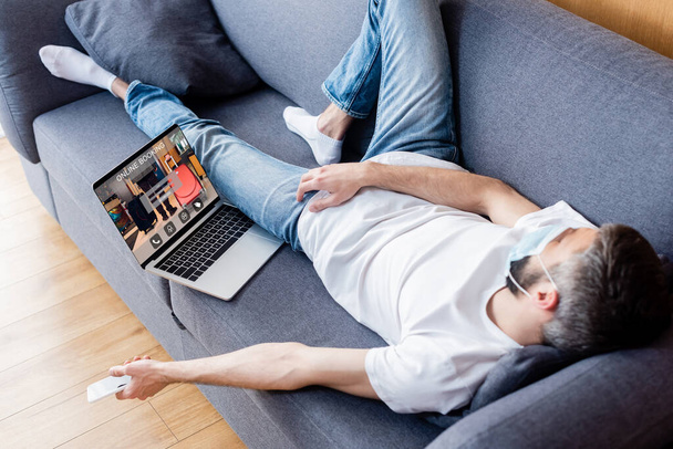 Hombre con máscara médica sosteniendo teléfono inteligente mientras duerme cerca de la computadora portátil con sitio web de reserva en línea en la pantalla
  - Foto, imagen