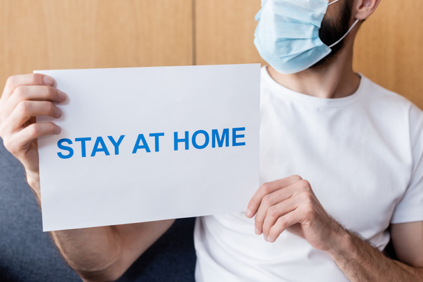 Cropped view of man in medical mask holding card with stay at home lettering on couch  - Photo, Image
