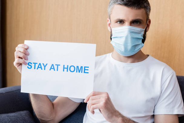 Man in medical mask holding card with stay at home lettering και κοιτάζοντας κάμερα στον καναπέ  - Φωτογραφία, εικόνα