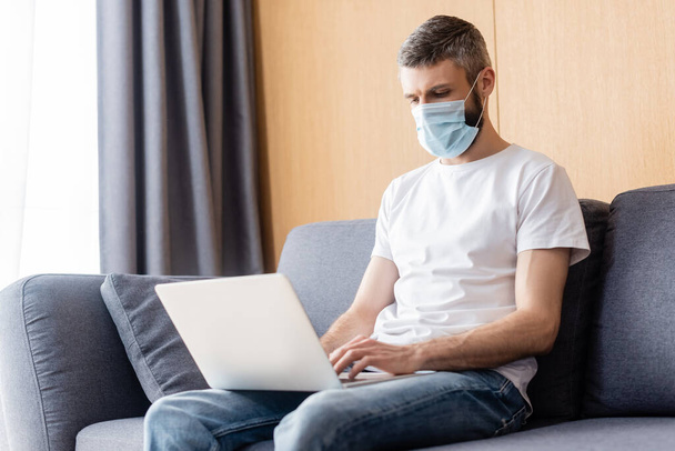 Teleworker in medical mask working on laptop on couch at home  - Φωτογραφία, εικόνα