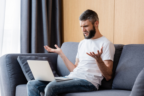 Confused freelancer looking at laptop while sitting on couch at home  - Photo, Image