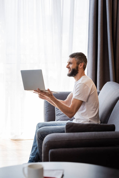 Side view of smiling freelancer having video chat on laptop in living room - Fotó, kép