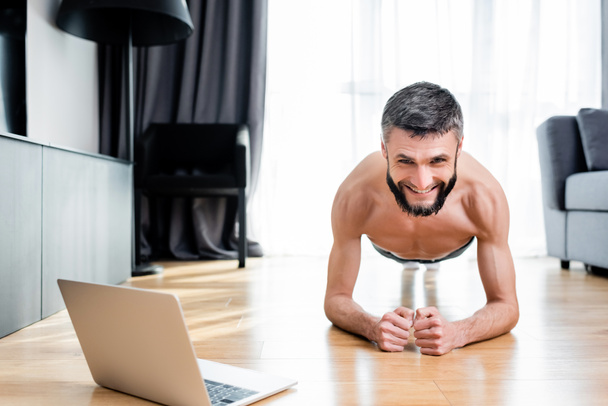 Shirtless man smiling at camera while standing in plank near laptop on floor at home  - Zdjęcie, obraz