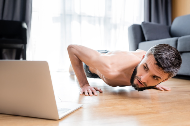 Selektiver Fokus des hemdslosen Sportlers, der Liegestütze in der Nähe des Laptops auf dem Boden macht  - Foto, Bild