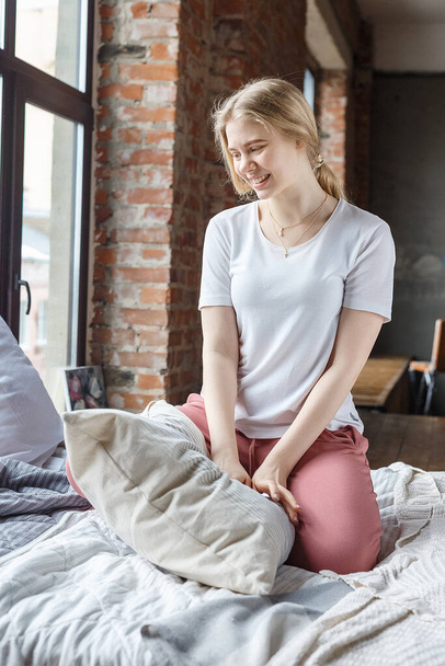 Blijf thuis concept: schattig blond jong mooi meisje in roze broek, wit t-shirt op bed in loft appartement glimlachend, met kussen in de armen. pyjamafeest tijdens COVID-19 pandemische zelfisolatie quarantaine - Foto, afbeelding