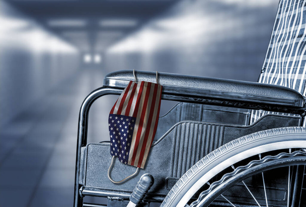 Flag of United States on face mask hanging on empty wheelchair in hallway of American hospital or retirement nursing care home with copy space. - Foto, afbeelding