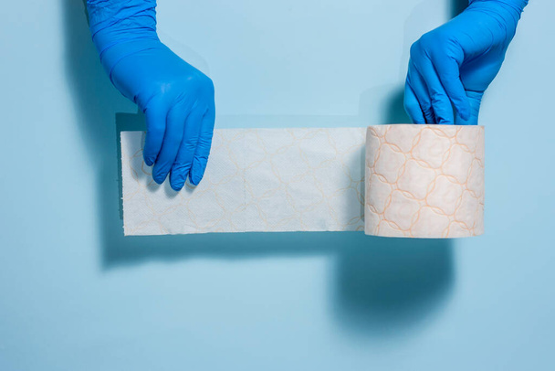 Dos manos con guantes de goma estériles sostienen un rollo de papel higiénico sobre un fondo azul. Símbolo de la pandemia del coronavirus
. - Foto, Imagen