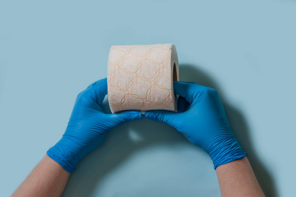 Dwie ręce w sterylnych gumowych rękawiczkach trzymają rolkę papieru toaletowego na niebieskim tle. Symbol pandemii koronawirusowej. - Zdjęcie, obraz