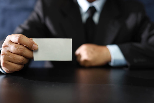Hombre de negocios manos en traje negro sentado y mostrando o dando tarjeta de nombre comercial en blanco mientras está sentado en la sala de reuniones. - Foto, Imagen