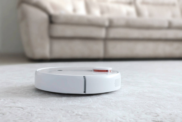 Aspirador robot blanco en alfombra. Moderno dispositivo inteligente piso de limpieza
. - Foto, Imagen