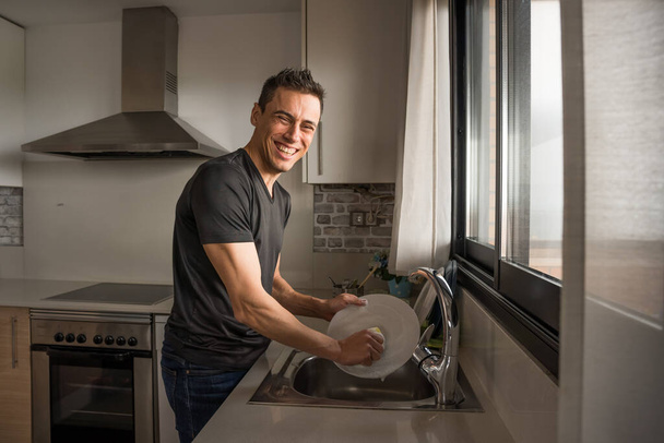 Улыбающийся и счастливый мужчина моет посуду на кухне рядом с окном. Средний выстрел
. - Фото, изображение