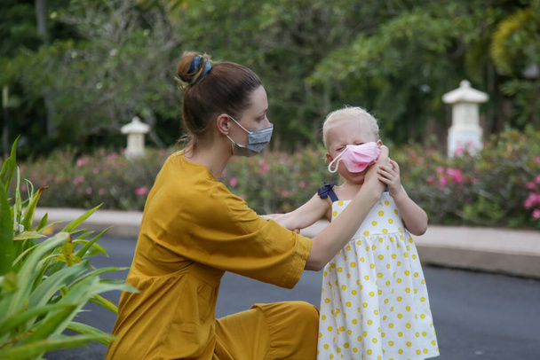 Mumie ringt darum, ihrem Kind eine medizinische Maske über das Gesicht zu ziehen. Rücksichtsloses Kleinkind weigert sich, einen Mundschutz zu tragen. Herausforderungen für Eltern bei Ausbruch der Coronavirus-Pandemie. - Foto, Bild