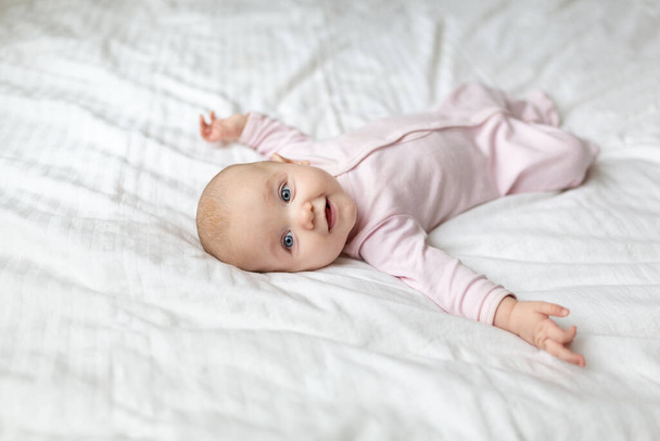 A little baby is smiling lying on the bed on a white sheet in the parents bedroom. A pink jumpsuit in newborn. Cute baby lying on the bed. Closeup portrait of newborn baby looking at camera - Foto, Imagem