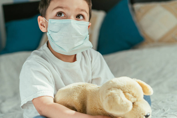 roztomilý batole chlapec v lékařské masce drží měkkou hračku doma - Fotografie, Obrázek