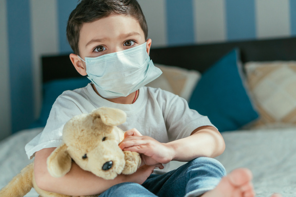 selective focus of toddler boy in medical mask holding soft toy at home - Foto, imagen