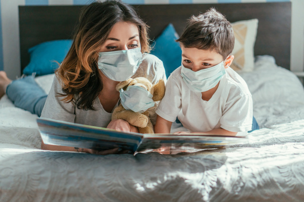 мать и ребенок сын в медицинских масках чтение книги возле мягкой игрушки
 - Фото, изображение