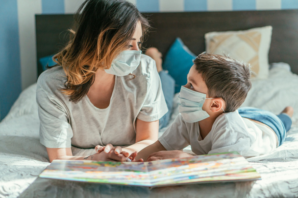 Mutter und Kleinkind-Sohn in Arztmasken schauen einander in der Nähe von Buch an  - Foto, Bild