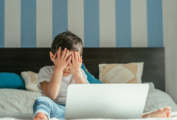 selective focus of toddler boy covering eyes while sitting near laptop in bedroom  - Fotó, kép
