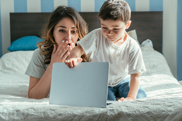 shocked freelancer covering mouth and looking at toddler son touching laptop in bedroom  - Foto, imagen