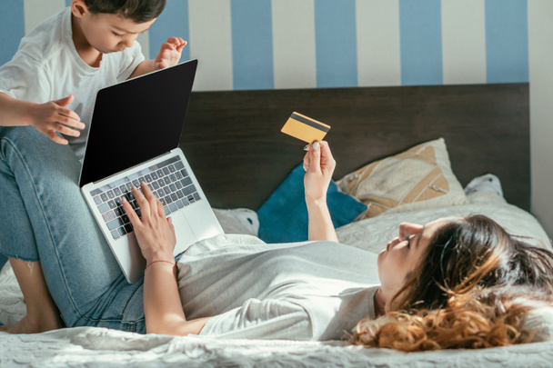 selective focus of woman lying on bed and holding credit card near laptop with blank screen and toddler son  - Photo, Image