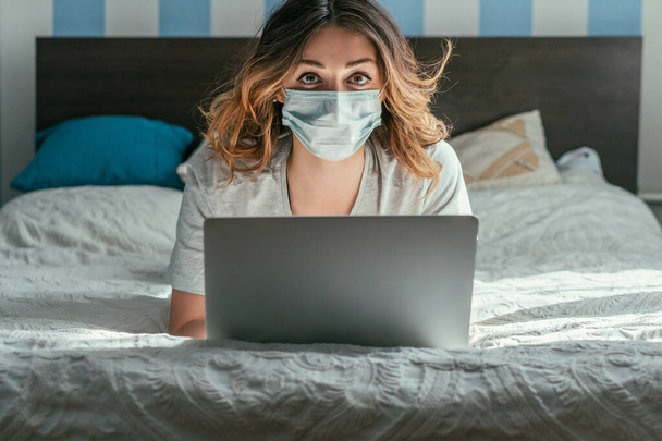 自宅で仕事をしながらベッドに寝そべってノートパソコンを使う医療用マスクのフリーランサー  - 写真・画像