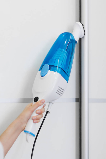 μια γυναίκα στα λευκά καθαρίζει το πάτωμα με μια ηλεκτρική σκούπα χεριών. Γενικός καθαρισμός του σπιτιού. Η νοικοκυρά τακτοποιεί τα πράγματα. υπηρεσίες καθαρισμού - Φωτογραφία, εικόνα