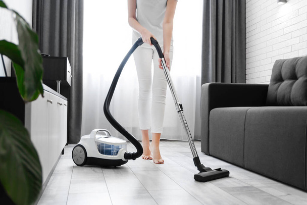 Μια γυναίκα στα λευκά καθαρίζει το πάτωμα με μια ηλεκτρική σκούπα. Γενικός καθαρισμός του σπιτιού. Η νοικοκυρά τακτοποιεί τα πράγματα. υπηρεσίες καθαρισμού - Φωτογραφία, εικόνα