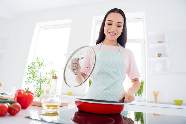 Photo of beautiful cheerful asian housewife hold frying pan checking meal condition cooking tasty dinner family wear t-shirt dotted apron stand modern kitchen indoors - Фото, зображення