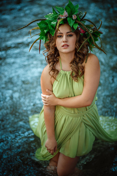 Όμορφη νεαρή γυναίκα με πράσινο φόρεμα και στεφάνι σε ένα ηλιόλουστο δάσος. τελετή στα μέσα του καλοκαιριού ή την Ημέρα της Γης. ιδέα και έννοια του αρώματος, παραμύθια και ευτυχία - Φωτογραφία, εικόνα