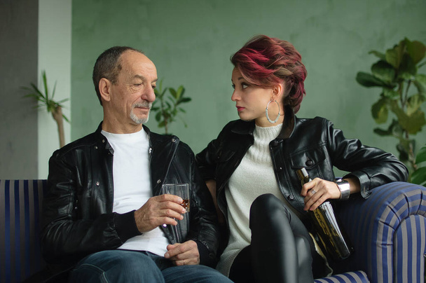 Senior Vater trinkt mit seiner erwachsenen Tochter auf blauem Sofa im Loft-Stil Zimmer, Mann hält Glas Whisky, Frau hält Flasche in der Hand, Alkoholsucht Konzept - Foto, Bild