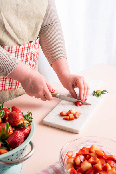 Onherkenbare huisvrouw snijdt de aardbeien in kleine stukjes. Het proces van het thuis maken van aardbeienjam met biologisch fruit. Vrouw bereidt de vruchten voor jam - Foto, afbeelding