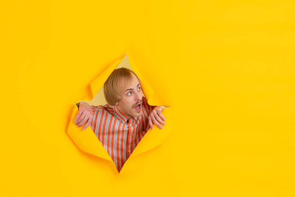 Cheerful young man poses in torn yellow paper hole background, emotional and expressive - Zdjęcie, obraz