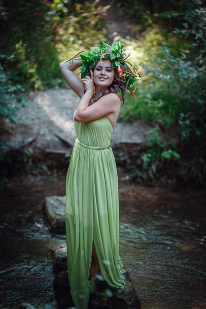 Mooie jonge vrouw in een groene jurk en een krans in een zonnig bos. ceremonie op midzomer- of aardedag. idee en concept van parfum, sprookjes en geluk - Foto, afbeelding
