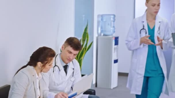 In the modern hospital corridor doctors have a discussing with the patient other two young doctors walking in front of the camera to the hospital reception discussing some diagnostic of the patient - Video