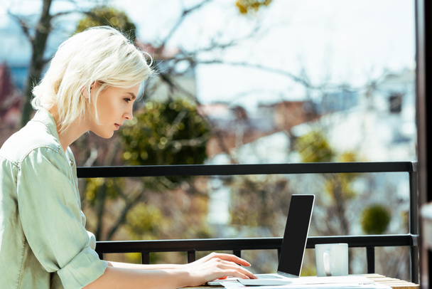 side view of blonde freelancer sitting on terrace and using laptop - 写真・画像