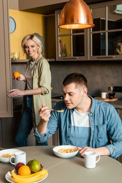 усміхнена жінка стоїть біля чоловіка, їсть кукурудзяні пластівці на кухні
 - Фото, зображення