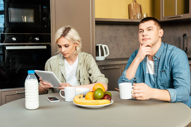 woman using digital tablet near dreamy husband while having breakfast in kitchen - Foto, afbeelding