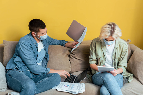 pareja joven en máscaras médicas en el sofá utilizando el ordenador portátil y la tableta digital cerca de papeles durante la cuarentena
 - Foto, imagen