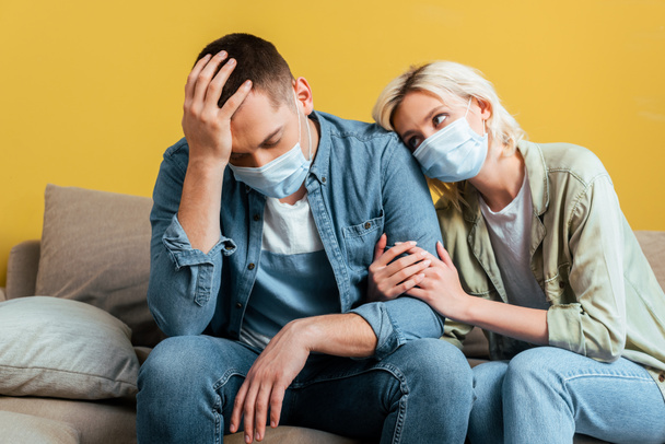 young sad couple in medical masks on sofa near yellow wall - Foto, Imagem