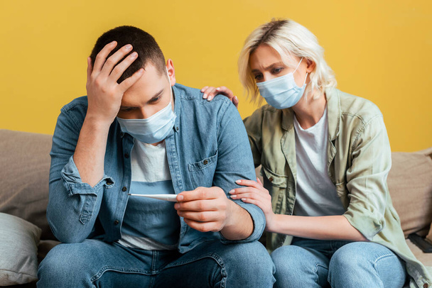 sad young couple in medical masks with thermometer on sofa near yellow wall - Foto, Bild