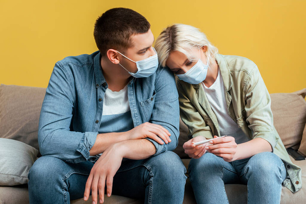 sad young couple in medical masks with thermometer on sofa near yellow wall - Photo, Image