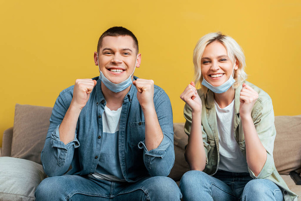 cheerful couple in medical masks showing yes gestures on sofa near yellow wall - Photo, Image