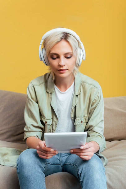 young blonde woman in headphones with digital tablet on sofa near yellow wall - Foto, imagen