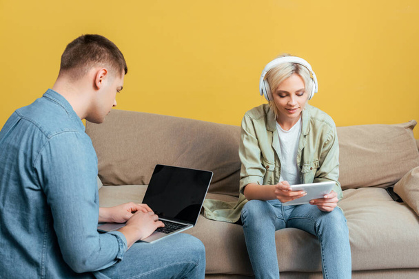 smiling young blonde woman in headphones with digital tablet near boyfriend with laptop on sofa near yellow wall - Foto, Imagem