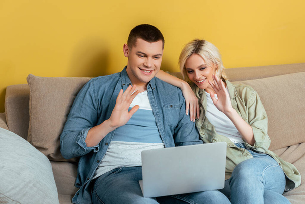 happy young couple on sofa  having video chat on laptop and waving hand near yellow wall - Φωτογραφία, εικόνα