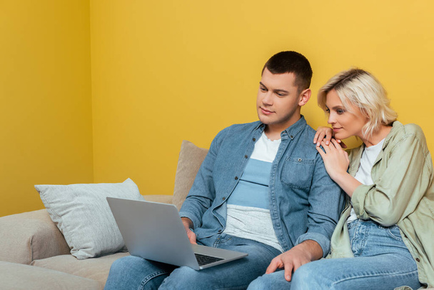 young couple on sofa sitting with laptop near yellow wall - Zdjęcie, obraz