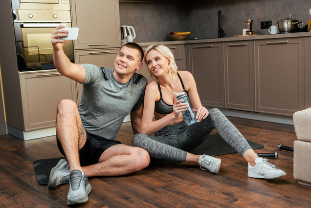 uśmiechnięta młoda para siedząca razem na macie fitness i robiąca selfie na smartfonie w domu podczas kwarantanny - Zdjęcie, obraz