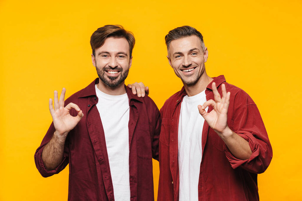 Image of two handsome men 30s in red shirts smiling and gesturing okay sign isolated over yellow background - Zdjęcie, obraz